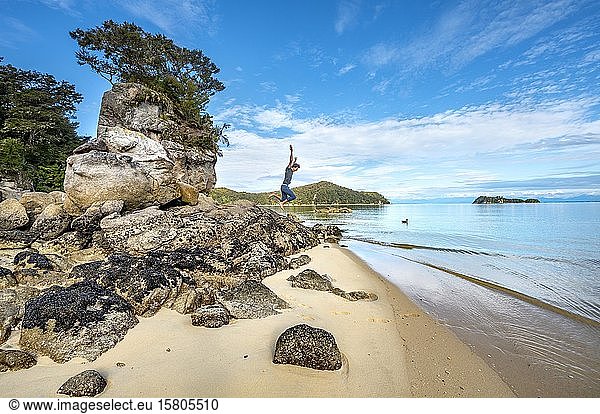 Junger Mann springt von einem überwucherten Felsen am Strand der Stillwell Bay  Abel Tasman National Park  Tasman  Südinsel  Neuseeland  Ozeanien
