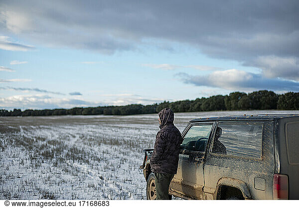Junger Mann steht neben einem Auto auf einem landwirtschaftlichen Feld im Winter