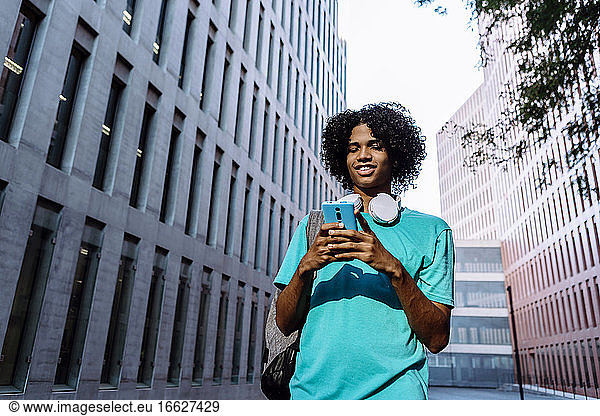 Junger Mann Textnachrichten auf Smartphone stehend gegen Gebäude außen in der Stadt