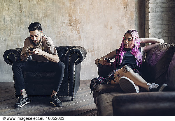 Junger Mann und Frau sitzen auf Sofa und Sessel in einem Loft und benutzen Smartphones
