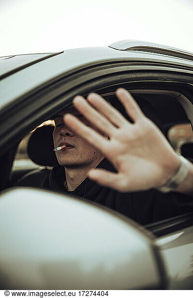 Junger Mann zeigt Stopp-Geste beim Rauchen einer Zigarette im Auto