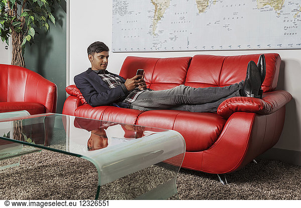Junger Millennial-Geschäftsmann benutzt sein Smartphone in der Lobby eines Arbeitsplatzes  während er auf einer Couch liegt; Sherwood Park  Alberta  Kanada