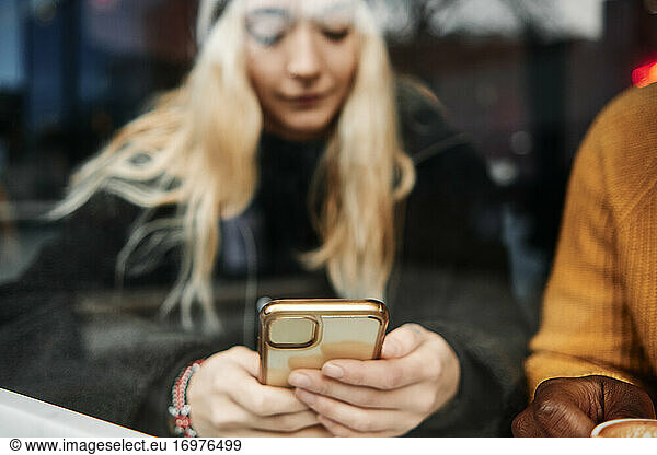 junges mädchen überprüft ihr telefon  am fenster eines cafés sitzend. spiegelung auf dem glas. fokus auf das telefon