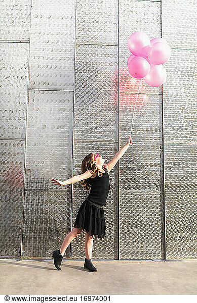 Junges Mädchen mit rosa Brille und rosa Luftballons vor einer Metallwand.