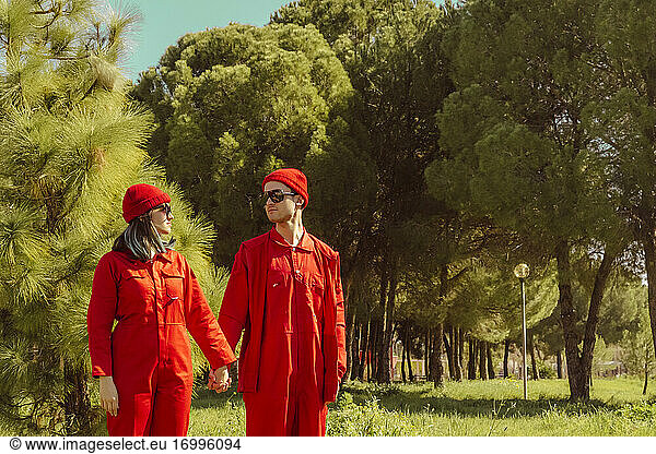Junges Paar in roten Overalls und Hüten steht Hand in Hand auf einer Wiese und sieht sich an