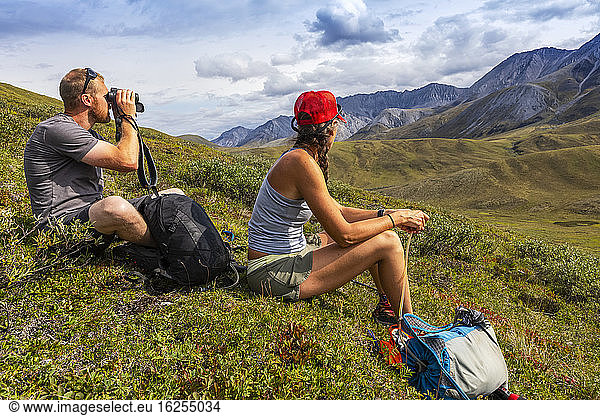 Kaukasischer Mann in den 30er Jahren  der sich hinsetzt und durch ein Fernglas nach Wildtieren schaut  während eine kaukasische Frau in den 40er Jahren neben ihm sitzt und die Berge in die gleiche Richtung schaut  während sie eine Wanderpause einlegt  Brooks Range; Alaska  Vereinigte Staaten von Amerika