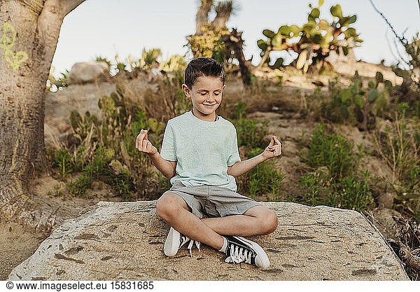 Kindergarten age boy meditating on rock in sunny cactus garden