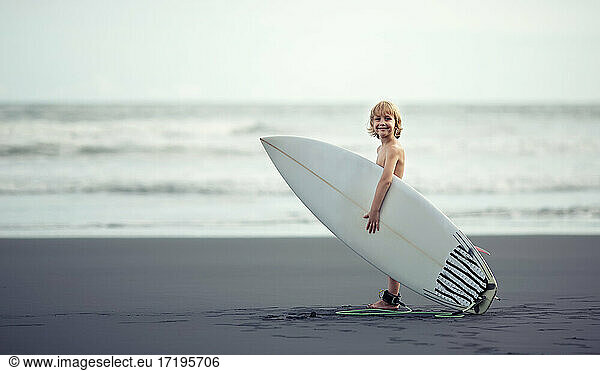 Kleiner Junge in der Nähe des Ozeans mit einem Surfbrett  der in die Kamera lächelt