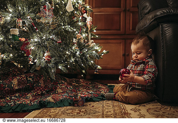 Kleiner Junge sitzt unter dem Weihnachtsbaum und hält ein Ornament