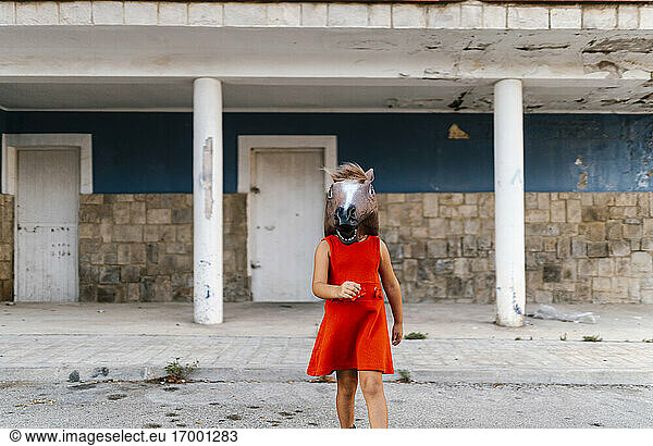 Kleines Mädchen mit einem Pferdekopf und einem roten Kleid  das von einem verlassenen Haus weggeht