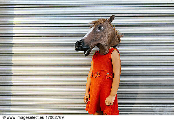 Kleines Mädchen mit einem Pferdekopf und einem roten Kleid vor einem Metalltor