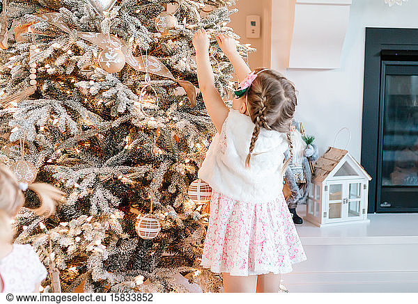 Kleinkind in rosa Kleid legt ein Ornament an einen beflockten Weihnachtsbaum
