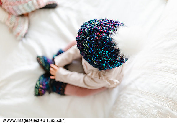 Kleinkind sitzt auf einem weißen Bett in einer Strickmütze mit weißem Bommel