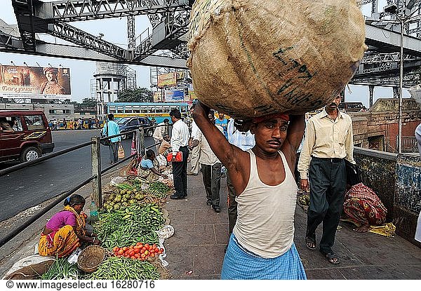 Kolkata (Kalkutta)  Westbengalen  Indien  Asien - Ein Kuli geht zusammen mit anderen Menschen an einem Straßenmarkt an einem Ende der Howrah-Brücke vorbei  an dem täglicher Verkehr vorbeifließt.
