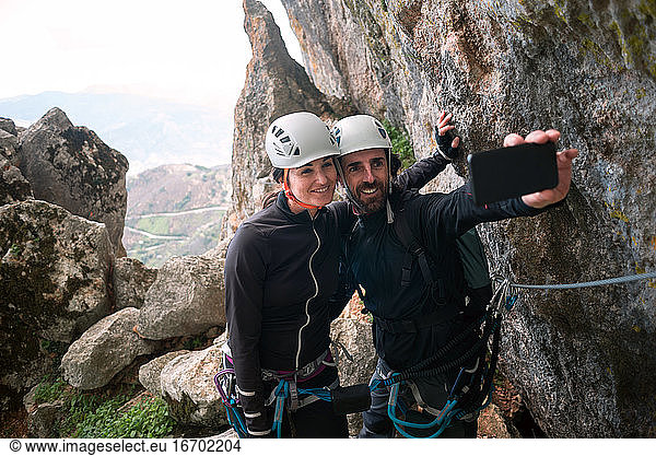 Konzept: Abenteuer. Ein Paar Bergsteiger mit Helm und Gurt. Sie machen ein Selfie mit dem Smartphone. Auf dem Gipfel des Berges. Via ferrata in Fels.