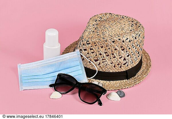 Konzept für Sommerurlaub während der Coronavirus Krise mit Gesichtsmaske  Strohhut  Sonnenbrille  Desinfektionsmittel Sprühflasche und Muscheln auf rosa Hintergrund