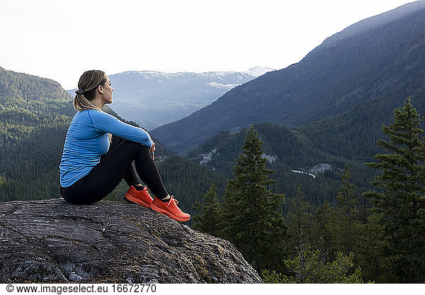 Kräftige Sportlerin  die beim Fitnesstraining auf dem Lande vor einem bewaldeten Bergrücken sitzt und eine Pause vom Trailrunning einlegt