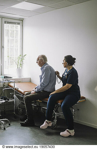 Krankenschwester mit Stethoskop bei der Untersuchung eines älteren männlichen Patienten  der auf einem Bett in einer Klinik sitzt