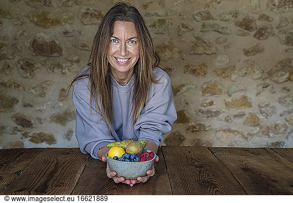 Lächelnde Ernährungsberaterin mit verschiedenen Früchten in einer Schale an einem Tisch vor einer Steinwand sitzend