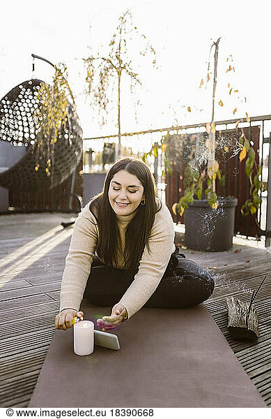 Lächelnde Frau  die eine Kerze anzündet  während sie auf einer Übungsmatte auf der Terrasse sitzt