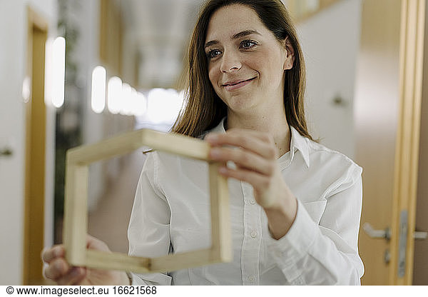 Lächelnde Frau  die einen Holzrahmen hält  während sie im Büro steht