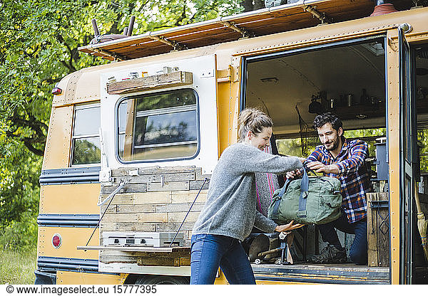 Lächelnde Frau hilft Mann beim Ausladen des Gepäcks aus gelbem Wohnmobil beim Camping im Wald