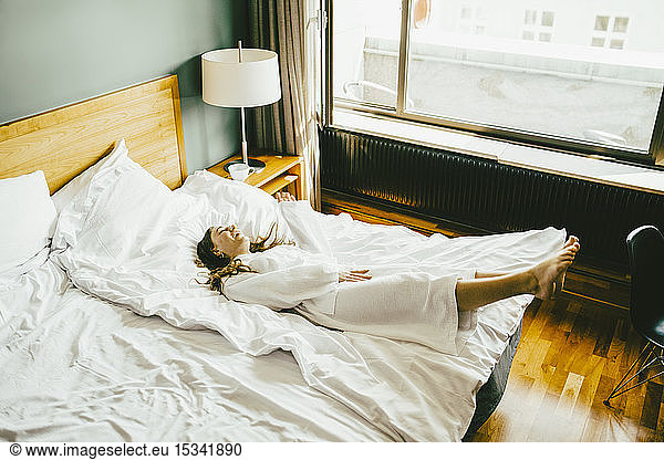 Lächelnde Frau im Bademantel amüsiert sich im Hotelzimmer im Bett