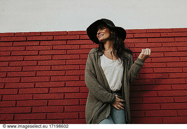 Lächelnde Frau in Strickjacke schaut weg  während sie an einer roten Backsteinmauer steht