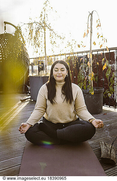 Lächelnde Frau macht Yoga im Schneidersitz auf einer Übungsmatte auf einer Terrasse