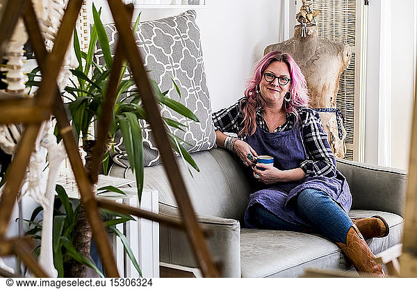 Lächelnde Frau mit langen blonden gewellten Haaren mit rosa Strähnen  die auf einem Sofa sitzt und in die Kamera schaut.