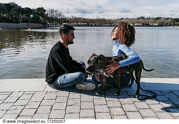 Lächelnde Freundin mit Freund sitzt mit Hund auf dem Fußweg am See