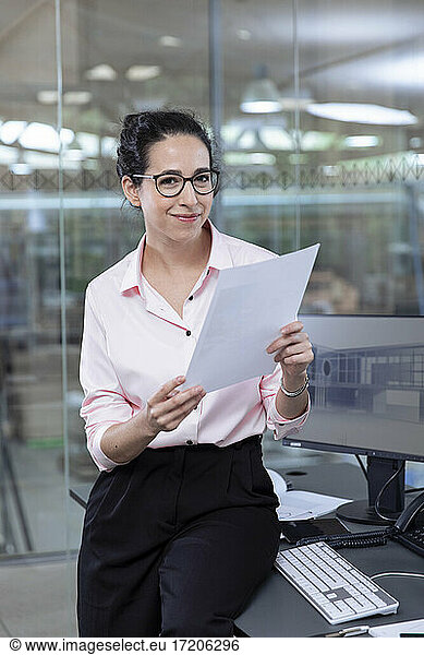 Lächelnde Geschäftsfrau mit Strategie sitzt am Schreibtisch im Büro