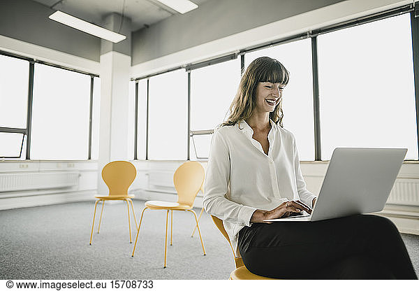 Lächelnde Geschäftsfrau sitzt mit einem Laptop auf einem Stuhl in einem leeren Büro