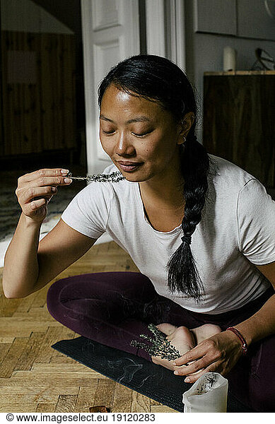 Lächelnde junge Frau  die an Kräutern riecht  während sie zu Hause auf einer Übungsmatte sitzt