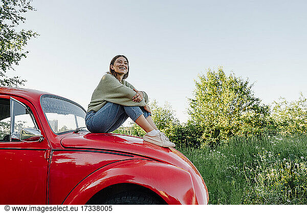 Lächelnde junge Frau  die auf der Motorhaube eines Autos sitzt und die Knie umarmt