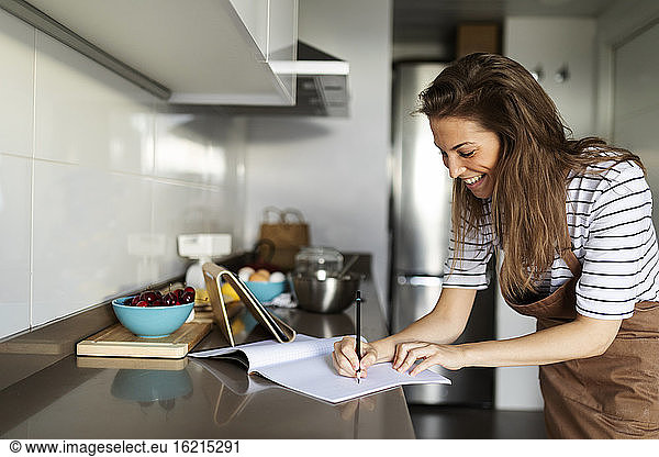 Lächelnde junge Frau  die ein Rezept in ein Buch schreibt  während sie zu Hause ein Video über ein digitales Tablet ansieht