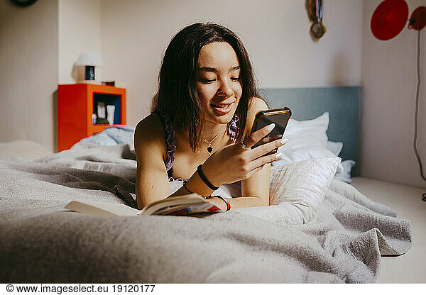 Lächelnde junge Frau  die ein Smartphone benutzt  während sie zu Hause auf dem Bett liegt