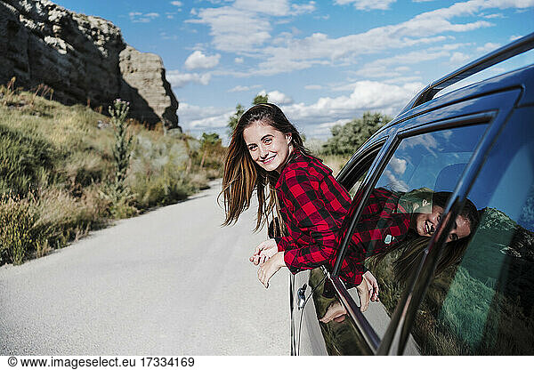 Lächelnde junge Frau lehnt sich aus dem Autofenster und genießt den sonnigen Tag