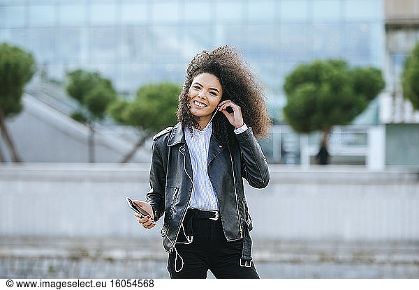 Lächelnde junge Frau mit Afrofrisur  die im Freien stehend Musik hört