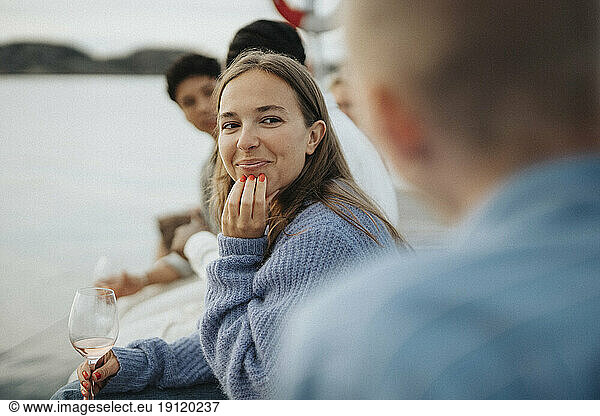 Lächelnde junge Frau mit Hand am Kinn  die mit Freunden auf einem Steg am See sitzt