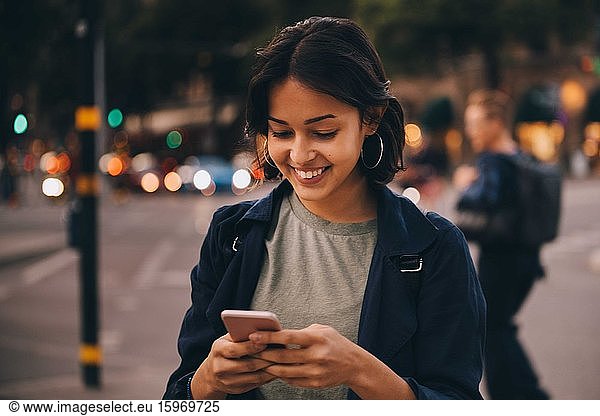 Lächelnde junge Frau nutzt soziale Medien am Telefon  während sie in der Stadt steht