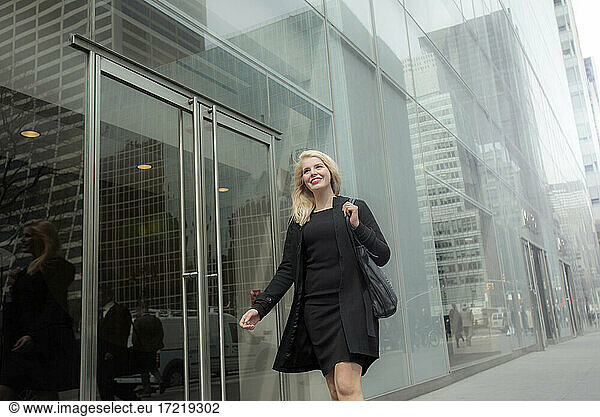 Lächelnde junge Geschäftsfrau geht an einem Glasgebäude vorbei