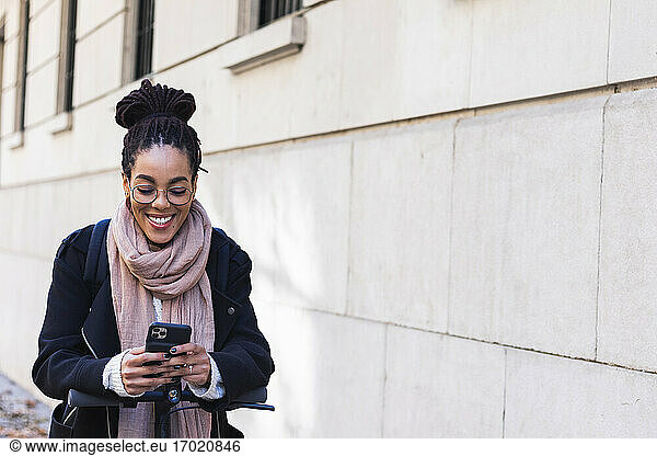 Lächelnde junge weibliche Millennials  die ihr Smartphone benutzen  während sie sich auf einen Elektroroller am Gebäude stützen