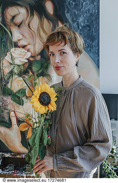 Lächelnde Künstlerin  die eine Sonnenblume hält und vor einem Kunstwerk steht