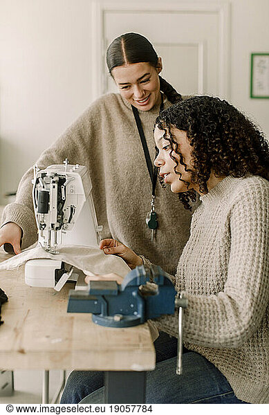 Lächelnde Lehrerin  die eine Schülerin bei der Arbeit an der Nähmaschine im Kunstunterricht der High School beobachtet