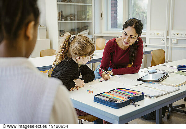 Lächelnde Lehrerin erklärt Schülerin am Schreibtisch sitzend im Klassenzimmer