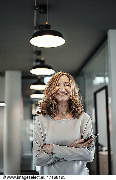 Lächelnde ältere Geschäftsfrau mit Smartphone in beleuchtetem Büro