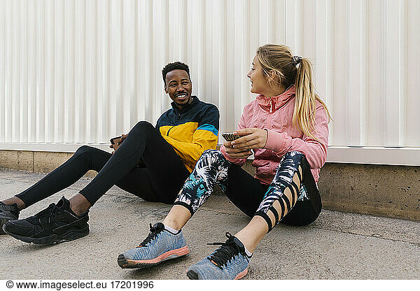 Lächelnde männliche und weibliche Athleten  die ein Mobiltelefon halten  während sie miteinander an der Wand sprechen
