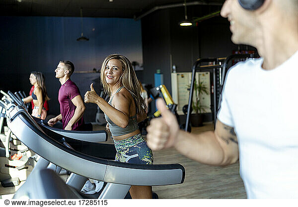 Lächelnde männliche und weibliche Athleten  die sich beim Training auf dem Laufband im Fitnessstudio gegenseitig die Daumen nach oben strecken