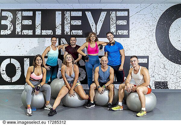 Lächelnde männliche und weibliche Athleten stehen hinter Freunden  die auf einem Fitnessball im Fitnessstudio sitzen